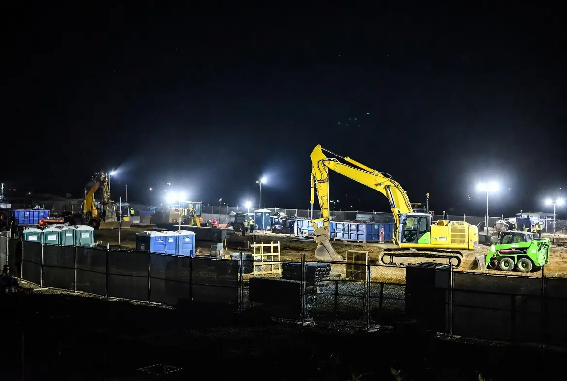 Nighttime construction jobsite in California illuminated by bright work lights, featuring temporary fencing, porta potties, excavator, skid steer, and workers in safety vests