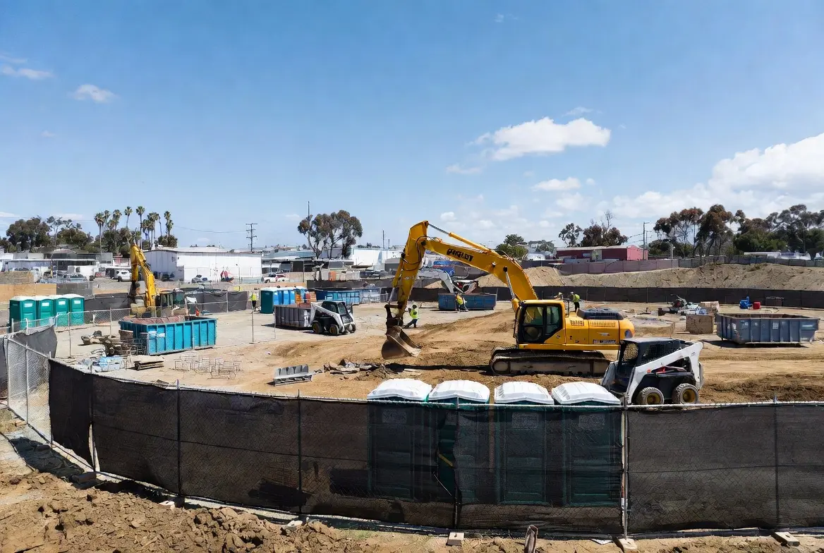 Busy construction jobsite in California with temporary fencing, porta potties, roll-off dumpsters, excavator, and skid steer loader working during the day